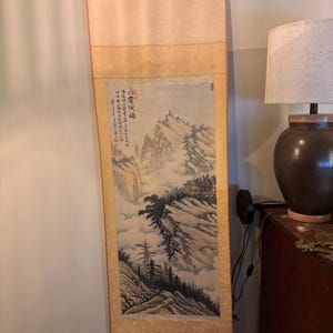 Peut inclure: Peinture sur rouleau verticale représentant un paysage montagneux avec du texte. L'œuvre utilise une palette de couleurs sourdes avec des nuances de beige, de gris et de vert. Le rouleau est encadré d'une bordure beige clair et possède des chevilles en bois en bas.