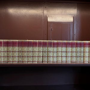 Pode incluir: Uma coleção de vinte e quatro livros de capa dura dispostos em fila. Cada livro apresenta uma lombada de cor creme com um padrão circular dourado repetitivo e uma faixa vermelha escura no topo, com linhas e letras douradas.