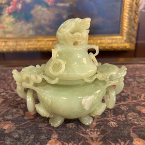 May include: A pale green carved stone incense burner featuring a lid topped with a mythical creature figure. The vessel has two side handles with dangling rings and is supported by three small feet. The surface shows smooth, polished textures.