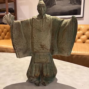 Op de afbeelding: Een bronzen beeld van een geklede figuur met uitgestrekte armen, die een waaier vasthoudt. Het beeld heeft ingewikkelde details op de gewaden en een puntmuts. Het gezicht van de figuur heeft een serene uitdrukking. Het beeld is een decoratief item.