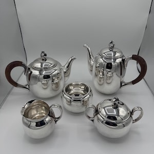 Puede incluir: Un juego de té de plata con tetera, lechera, azucarero y dos piezas de servicio adicionales. La tetera tiene un asa marrón oscuro. El juego es muy reflectante, con la luz reflejada en las superficies pulidas.