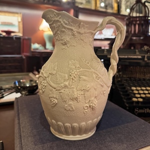 Puede incluir: Jarra de cerámica color crema con superficie texturizada y diseño en relieve de uvas y vides. El asa está intrincadamente torcida. La jarra está colocada sobre un libro azul, lo que sugiere una estética vintage o antigua.