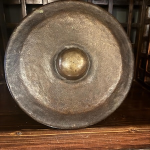May include: A bronze gong with a central raised boss. The circular percussion instrument has a dark, aged patina and sits on a wooden surface. The gong's surface shows signs of wear, suggesting age and use. The background includes a wooden structure.