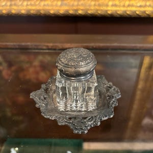 May include: An antique inkwell with a clear glass ink pot and a decorative silver-toned lid. The inkwell sits on a matching ornate silver-toned base. The inkwell is displayed on a glass surface, with a blurred background.