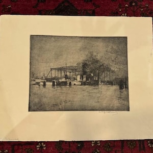 Op de afbeelding: Een zwart-wit ets van een waterkantscène, met gebouwen en structuren langs de waterkant. Het kunstwerk is ingelijst in een witte passe-partout, met de handtekening van de kunstenaar zichtbaar.