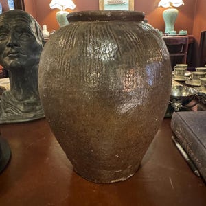 May include: A large, brown, glazed ceramic jar with a wide, rounded body and a narrow neck. The jar has vertical textured lines and sits on a dark wooden surface. The jar is a decorative piece.