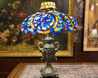 Op de afbeelding: Een tafellamp met een glas-in-lood kap met blauwe, groene en rode bloemmotieven. De voet is gemaakt van donker, bewerkt metaal met twee handvatten aan de zijkanten en een vierkante, gedetailleerde voet. Twee trekkoorden hangen aan de fitting.