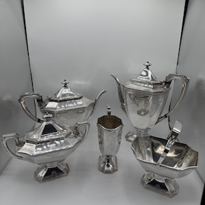 Puede incluir: Un juego de té plateado con detalles ornamentados. El conjunto incluye una tetera, azucarero, cremera y un cuenco para servir con pinzas. Las piezas tienen una superficie reflectante y están dispuestas sobre un fondo blanco.