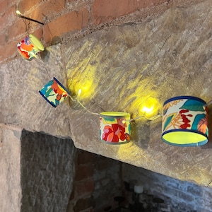 May include: A string of fairy lights with colourful floral patterned paper lampshades hanging from a brick wall. The lights are turned on and glowing.