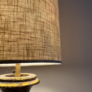 May include: A table lamp featuring a cylindrical lampshade with a textured, cross-hatched beige fabric pattern. The base is constructed from a dark, weathered material with visible yellow paint distressing along the edges and a brass-colored neck.