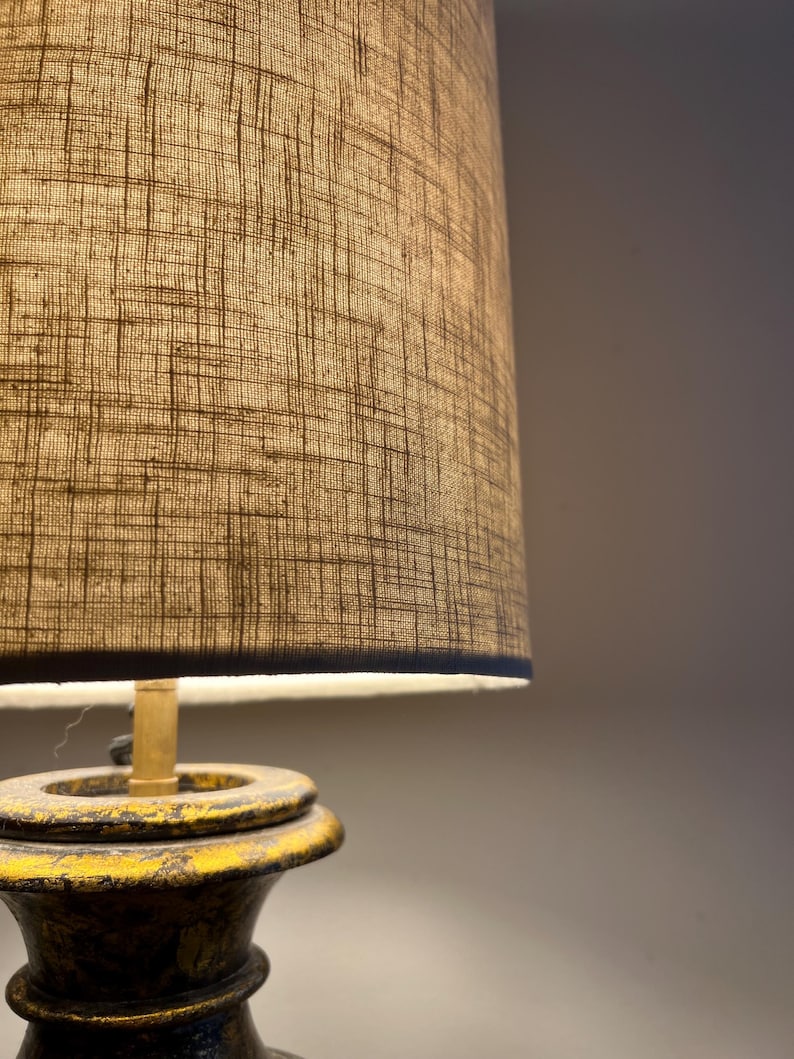May include: A table lamp featuring a cylindrical lampshade with a textured, cross-hatched beige fabric pattern. The base is constructed from a dark, weathered material with visible yellow paint distressing along the edges and a brass-colored neck.