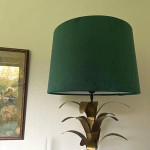 May include: A green lampshade sits atop a gold-colored lamp base with a palm frond design. The lampshade is a deep green color and has a conical shape. The lamp is placed in front of a white wall, with a framed picture on the left.