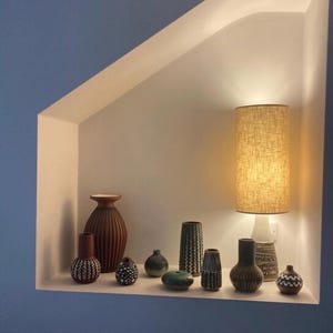 May include: A collection of ten ceramic vases of varying shapes, sizes, and textures, featuring earthy tones of brown, green, and cream, arranged on a shelf alongside a tall cylindrical table lamp with a textured beige fabric shade.