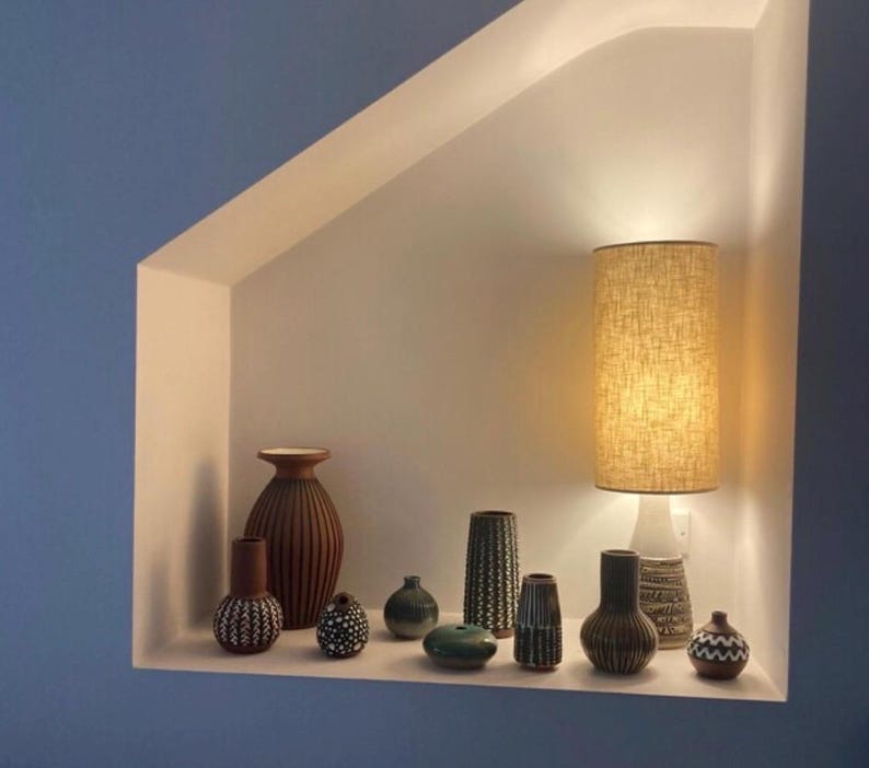 May include: A collection of ten ceramic vases of varying shapes, sizes, and textures, featuring earthy tones of brown, green, and cream, arranged on a shelf alongside a tall cylindrical table lamp with a textured beige fabric shade.
