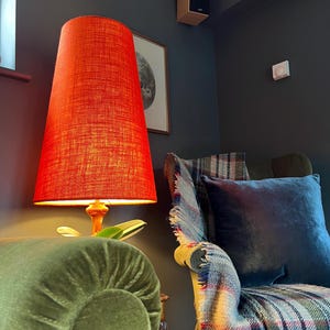 May include: A tall, orange lampshade with a textured surface sits atop a wooden lamp base. The lamp is placed next to a green velvet armchair with a blue pillow and a plaid blanket. A framed picture hangs on the dark gray wall.