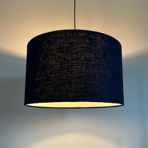 May include: A black, cylindrical lampshade hangs from the ceiling. The lampshade is made of a textured, dark fabric. The interior of the lampshade emits a warm, soft light. This modern pendant light is suitable for various interior design styles.