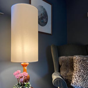 May include: A tall floor lamp with a cylindrical off-white fabric shade and a swirling orange and white glass base. Positioned in front of the lamp is a small potted plant with vibrant pink cyclamen flowers and green leaves.