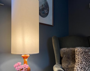 May include: A tall floor lamp with a cylindrical off-white fabric shade and a swirling orange and white glass base. The lamp is positioned next to a potted plant with pink flowers, a dark armchair with a faux fur pillow, and a framed wall picture.