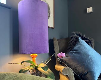 May include: A purple velvet lampshade with an orange base sits on a table next to a green armchair. The armchair has a blue velvet cushion with a black fur trim and a pink orchid.