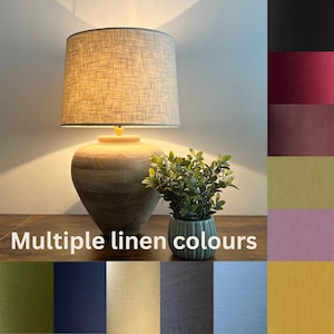 May include: A table lamp with a light-colored, textured linen drum shade and a rounded wooden base, illuminated to cast a warm glow. Beside it sits a small green plant in a ribbed pot. The text Multiple linen colours is visible.
