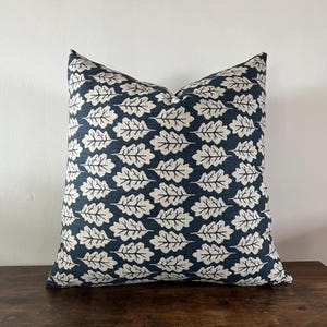 May include: A square throw cushion featuring a dark navy blue background with a repeating pattern of off-white oak leaves. The fabric has a visible woven texture, and the leaves are arranged in horizontal rows across the entire surface of the cushion.