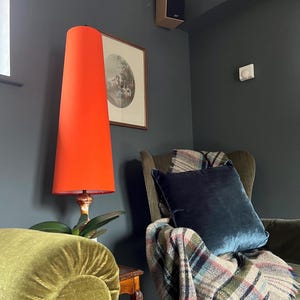 May include: A vibrant orange lampshade with a conical shape dominates the scene, set against a dark grey wall. The lamp has a decorative base. A framed picture hangs nearby, and a blue cushion rests on a patterned throw in a green armchair.
