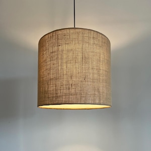 May include: A cylindrical lampshade made from a light brown, woven fabric suspended from a black cord. The lampshade is lit from within, producing a warm light. The neutral colour and simple design suit various interior styles.
