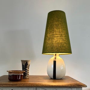 May include: A table lamp with a tall, tapered olive green fabric shade and a rounded white ceramic base featuring a vertical black stripe. Beside the lamp sits a small brown ceramic bowl and a tall, cylindrical vase with a black leaf pattern.