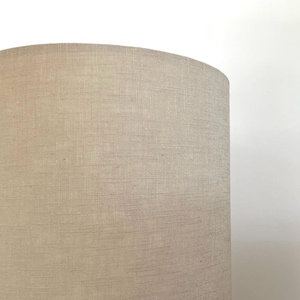 May include: A cylindrical lamp shade featuring a light beige, neutral-toned fabric with a visible cross-hatched, woven texture. The shade has a smooth, rounded top edge and a consistent, solid color throughout its exterior surface.