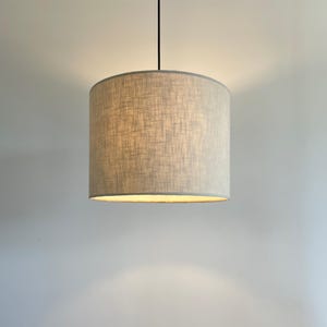 May include: A cylindrical pendant light fitting featuring a light grey fabric shade with a visible cross-hatch weave texture. The drum-shaped shade is suspended from a thin black cord and emits a warm, soft glow from the bottom opening.