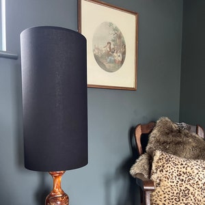 May include: A tall, black lamp with a cylindrical shade stands next to a wall. The lamp base has a marbled orange and brown pattern. A framed print with an oval image hangs on the wall, and a chair with a leopard print pillow is in the background.