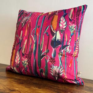 May include: A square throw cushion featuring a vibrant magenta background with a dense, colourful botanical print of various tropical flowers, tall stalks, and exotic plant life in shades of green, yellow, orange, and deep purple.