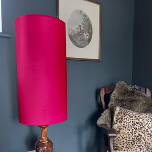 May include: A tall, cylindrical lamp shade in a vibrant magenta fabric. The shade is mounted on a polished, dark brown wooden base with a carved, swirling design. The lamp stands against a dark gray wall, positioned near a wooden chair with fur.