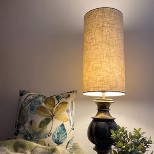 May include: A table lamp with a tall cylindrical beige fabric shade and a dark, weathered black and gold pedestal base. Beside it sits a square throw pillow featuring a botanical leaf print in shades of green, yellow, and blue.