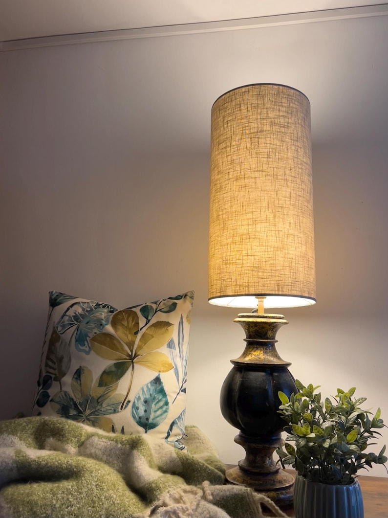 May include: A table lamp with a tall cylindrical beige fabric shade and a dark, weathered black and gold pedestal base. Beside it sits a square throw pillow featuring a botanical leaf print in shades of green, yellow, and blue.