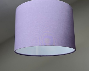 May include: A cylindrical lampshade in a soft lavender color hangs from a white cord. The lampshade has a smooth, fabric-like texture and a white interior. The light fixture is suspended from the ceiling.