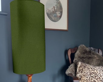 May include: A green fabric lampshade with a wooden base. The lampshade is cylindrical and has a textured surface. The lamp is sitting on a table in a room with a dark blue wall.