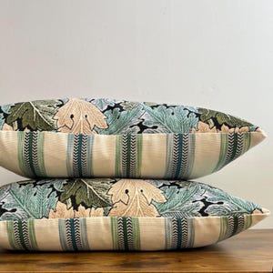 May include: Two rectangular throw pillows stacked vertically. The top half of each pillow features a woven botanical pattern with green, teal, and beige leaves on a black background. The bottom half features vertical stripes in cream, green, and teal.