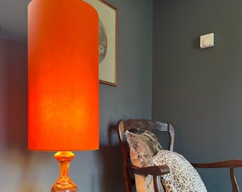 May include: A bright orange lamp with a cylindrical shade sits on a table in a room with a dark gray wall. The lamp has a marble base and a warm glow. A wooden chair with a leopard print cushion is in the background.