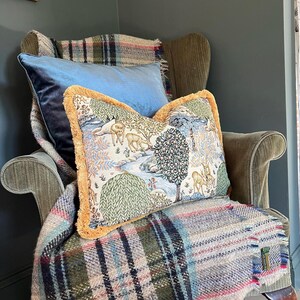 May include: A cozy armchair with a blue velvet pillow and a decorative pillow featuring a landscape scene with deer and trees. A plaid blanket with fringe is draped over the chair, creating a warm and inviting atmosphere.