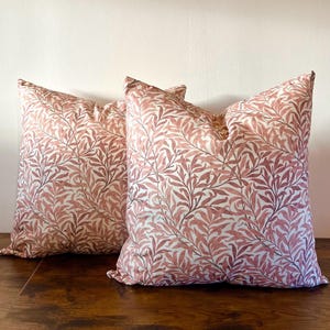 May include: Two square throw pillows with a botanical pattern. The pillows feature a repeating design of reddish-brown leaves and stems against a cream-colored background. The pillows are placed on a dark wooden surface.