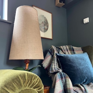 May include: A tall, beige burlap lampshade with a wooden base stands next to a green velvet armchair. A blue velvet pillow and a plaid blanket rest on the chair. A framed picture hangs on the dark blue wall, and a speaker is mounted on the ceiling.