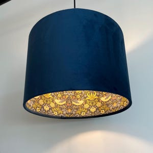 May include: A cylindrical pendant lampshade featuring a solid dark navy blue velvet exterior. The interior lining displays a detailed botanical pattern with yellow birds, flowers, and foliage set against a dark brown background, illuminated from within.