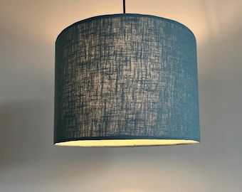 May include: A cylindrical pendant lamp shade featuring a textured, cross-hatched fabric pattern in a deep teal blue color. The shade is suspended by a thin black cord and emits a soft, warm glow from within, illuminating the fabric's weave.