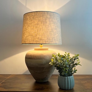May include: A table lamp with a light brown, horizontally banded wooden base and a cylindrical, textured beige fabric lampshade. The lamp emits a warm glow, casting light upwards and downwards. Beside it sits a small, ribbed, light green pot.