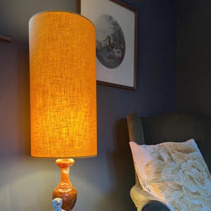 May include: A tall cylindrical lamp with a textured mustard yellow fabric shade and a polished orange and brown marbled stone base. The lamp emits a warm glow, illuminating the surrounding area with a soft light, standing next to a dark armchair.