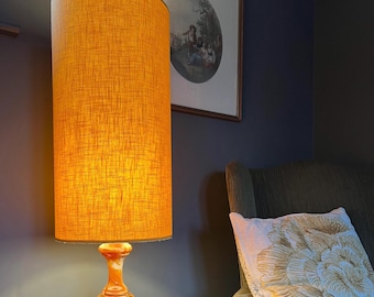 May include: A tall cylindrical lamp with a textured mustard yellow fabric shade and a polished orange and brown marbled stone base. The lamp emits a warm glow, illuminating the surrounding area with a soft light, standing next to a dark armchair.