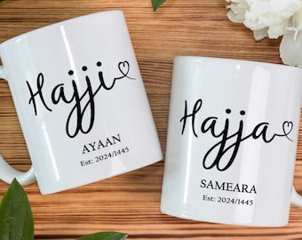 May include: Two white ceramic mugs with black calligraphy that reads "Hajji" and "Hajja" with a small heart symbol. The mug on the left has the name "Ayaan" and the date "Est. 2024/1445" printed below the calligraphy. The mug on the right has the name "Sameara" and the date "Est. 2024/1445" printed below the calligraphy.