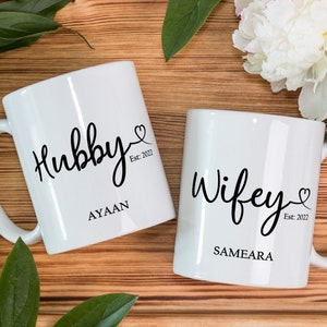 May include: Two white ceramic mugs with black lettering. One mug says "Hubby Est. 2022 Ayaan" and the other mug says "Wifey Est. 2022 Sameara".
