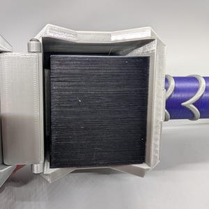 May include: A close-up of a 3D-printed object with a silver-gray frame and a black, ridged interior. The object has a purple and silver handle. The design appears to be a stylized weapon or tool.
